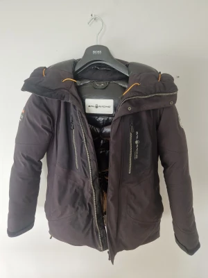 Svart Sail Racing dunjacka Gore-Tex - Svart dunjacka från Sail Racing med Gore-Tex-material, perfekt för kalla dagar. Jackan har huva med orange snörning, två bröstfickor med dragkedja och snygga logodetaljer på bröstet och ärmen. Riktigt schysst och funktionell design med robusta dragkedjor.