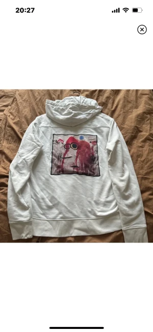 Vit hoodie med konsttryck från limitato - Vit hoodie från limitation  med ett stort konstnärligt tryck i rött, svart och blått på ryggen. Tröjan har huva och långa ärmar, tillverkad i mjuk bomull. Trycket har en unik, abstrakt design med ansiktsmotiv och färgstänk. Perfekt för dig som gillar streetwear med en twist.