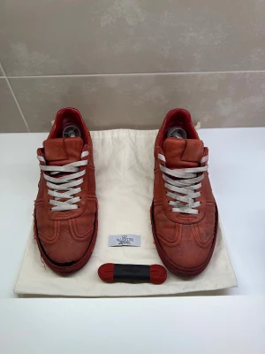 Valentino Garavani röda sneakers  - Säljer ett par röda Valentino Garavani sneakers i skinn med vita snören och extra röda skosnören. Snygg design med rund tå, platt sula och små nitar baktill på sulan. Perfekta för dig som gillar exklusiva och unika sneakers med streetkänsla.