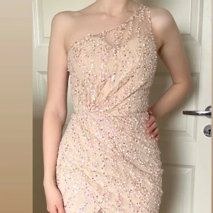 Champagnefärgad paljettklänning Jadore - Superfin långklänning från Jadore i champagnefärg med glittriga paljetter över hela klänningen. One shoulder-modell med slits framtill och figurnära passform. Perfekt för bal eller fest, ger en lyxig och elegant känsla. Helt ny och oanvänd!