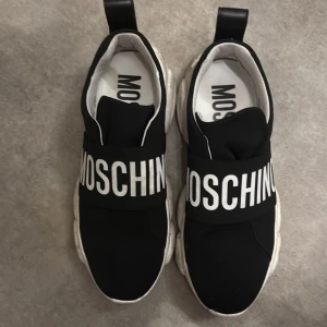 Svarta sneakers från Moschino - Säljer ett par svarta sneakers från Moschino med bred resår över foten och stor vit logga. Skorna har chunky vit sula med unik struktur och är tillverkade i syntetmaterial. Insidan är vit och de har en modern, sportig look som sticker ut. Billigare vid upphämtning 