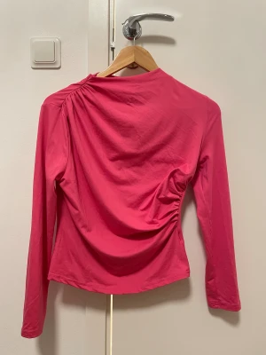 Rosa långärmad topp med drapering - Säljer en snygg rosa långärmad topp med cool drapering och rynkade detaljer på ena sidan. Toppen har en rund hals och är gjord i mjukt material som känns skönt mot huden. Perfekt för dig som vill ha en färgstark och unik look.