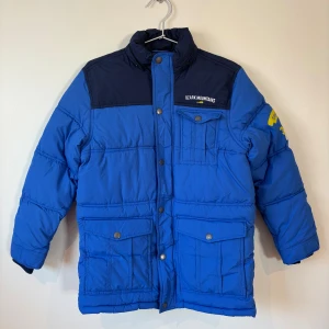  L.O.G.G. pufferjacka - 158 Storlek  - Blå och mörkblå pufferjacka från L.O.G.G. med trycket tryck på bröstet. Jackan har flera fickor med tryckknappar, hög krage och långa ärmar. Materialet är polyester och jackan är perfekt för kalla dagar.