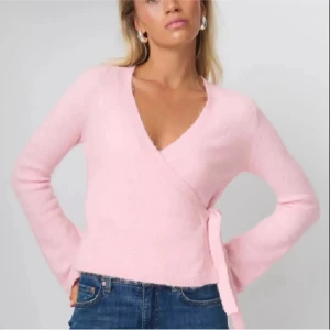 Ljusrosa omlottkofta från Gina Tricot - Superfin ljusrosa omlottkofta från Gina Tricot med knyt i sidan och v-ringad hals. Koftan är stickad och har långa ärmar, perfekt att slänga över en topp eller linne. Snygg och enkel att styla till jeans eller kjol.