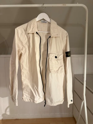 Vit overshirt från Stone Island - Snygg vit overshirt från Stone Island med klassisk patch på ärmen. Jackan har dragkedja framtill, stor bröstficka med knapp och elastisk nederkant. Tillverkad i bomull och perfekt för våren eller en julklapp till någon. Liten fläck ibak men ingenting man tänker på. Säljer pga att jag inte använder den, såklart äkta.