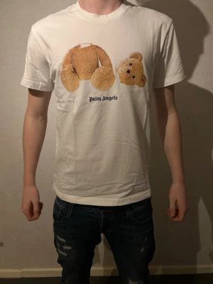 Vit Palm Angels t-shirt med nalletryck - Vit t-shirt från Palm Angels med ikoniskt tryck av en nallebjörn med avtagbart huvud på bröstet. Klassisk passform och rund halsringning. Tillverkad i mjuk bomull som känns skön mot huden. 