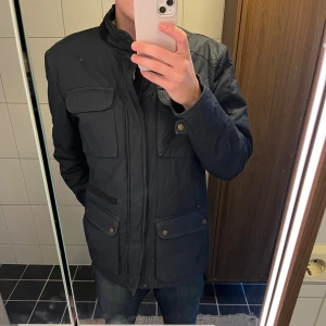 Svart parkajacka från Dressmann - Svart parkajacka från Dressmann i storlek L med flera fickor framtill och snygga detaljer som knappar och dragkedja. Jackan har hög krage och quiltad baksida, perfekt för kyliga dagar. Materialet är syntet och jackan är långärmad med normal passform.