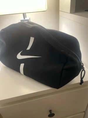 Svart träningsväska från Nike - Snygg svart träningsväska från Nike med stort vitt swoosh-tryck på sidan. Väskan har en robust dragkedja, rymligt huvudfack och är tillverkad i slitstark polyester. Perfekt för gymmet eller sportaktiviteter. Enkel och stilren design med diskret Nike-märke på baksidan.