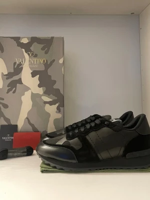 Valentino Garavani Rockrunner sneakers - Snygga svarta Valentino Garavani Rockrunner sneakers med coolt camouflage-mönster. Skorna har snörning, platt sula och detaljer i både skinn och mocka. Perfekt för dig som vill ha en exklusiv och trendig look med italiensk design.