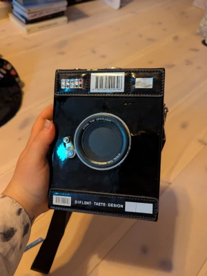 Svart axelväska i kameramodell - Unik svart axelväska formad som en retro kamera med coola detaljer som lins, knappar och silvriga metalldetaljer. Väskan har axelrem och är tillverkad i glansigt syntetmaterial. Perfekt för dig som vill sticka ut med en kreativ accessoar.