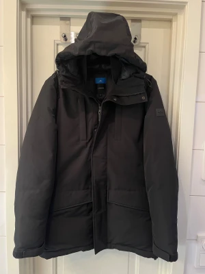 Svart dunjacka från J.Lindeberg - Säljer en svart dunjacka från J.Lindeberg med huva och snygga detaljer som dragkedja och knappar. Jackan har quiltad insida, justerbar huva med snörning och två stora fickor framtill. Perfekt för kalla vinterdagar och riktigt stilren look. • Mått📏 • Längd: 82cm • Bröstbredd: 55cm • Axelbredd: 48cm • Ärmlängd: 70cm • (Modellen på bilden är 178cm)  Nypris: 6000kr