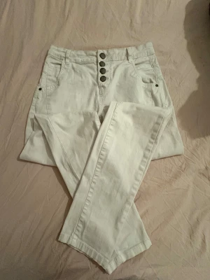 Vita highwaist jeans från KappAhl - Snygga vita jeans från KappAhl i storlek 34. Modellen är högmidjad med raka ben och har fem dekorativa knappar framtill. Byxorna är tillverkade i bomull med lite stretch för extra komfort. Perfekta för en fräsch och stilren look.