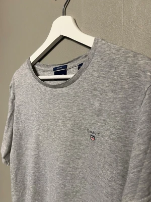 Grå t-shirt från GANT med logga - Snygg gråmelerad t-shirt från GANT med rund hals och diskret broderad logga på bröstet. Klassisk passform och mjukt bomullsmaterial som känns skönt mot huden. Perfekt för dig som gillar stilrena och enkla plagg.