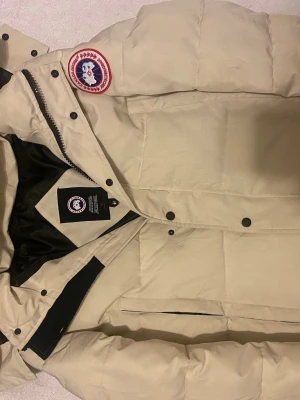 Beige Canada Goose dunjacka M - Säljer en beige dunjacka från Canada Goose i storlek M. Jackan har klassisk patch på ärmen, svarta knappar och dragkedja framtill. Fodret är svart och jackan har en stilren, boxig passform som håller dig varm under kalla dagar.