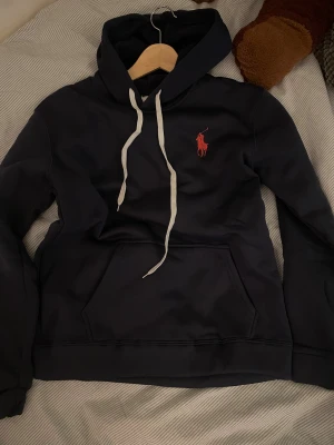 Mörkblå hoodie  - Snygg mörkblå hoodie  med klassisk röd logga på bröstet. Tröjan har vit snörning i huvan och en stor magficka framtill. Perfekt för dig som gillar stilren streetwear och vill ha en bekväm tröja med premiumkänsla.