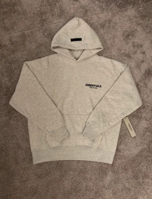 Grå Essentials Fear of God hoodie - Snygg äkta ljusgrå hoodie från Essentials Fear of God med svart logga på bröstet och stor tryckt logga på ryggen. Köpt ifrån farfetch för 3380kr. Perfekt för en clean och stilren streetwear-look. Priset kan diskuteras 