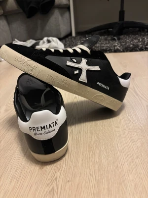 Premiata sneakers svart/vit skinn - Snygga Premiata sneakers i svart och vitt med kontrasterande logga på sidan och textdetaljer på hälen. Skorna har vit platt sula med mönstrad text, rund tå och vita snören. Tillverkade i skinn med stilren design och coola detaljer som sticker ut.