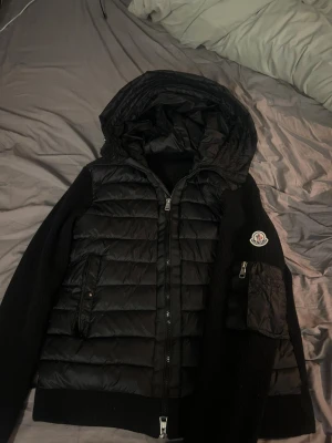 Svart Moncler pufferjacka med huva - Svart pufferjacka från Moncler med stickade ärmar och quiltad front. Jackan har huva, dragkedja framtill och en ficka med dragkedja på ärmen. Moncler-logga på vänster ärm. Snygg, modern look med blandat material för extra stilpoäng.