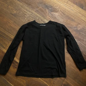 Svart långärmad topp från Calvin Klein - Svart långärmad topp från Calvin Klein Jeans i mjuk bomull. Enkel och stilren design med rund halsringning och diskret logga i nacken. Perfekt att matcha med jeans eller kjol för en clean look. Ny pris 499kr