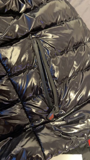 Svart Moncler pufferjacka L - Snygg svart pufferjacka från Moncler i storlek L. Jackan har glansig yta, quiltad design och dragkedja framtill samt på fickorna. Insidan har coolt Moncler-tryck och en röd detalj på innerfickan. Perfekt för dig som vill ha en stilren och varm jacka. Liten i storlek 