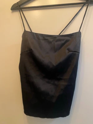 Svart satinlinnetopp med knytband - Säljer en svart linne-topp i satin med tunna axelband och snygg knytning i ryggen. Toppen har en öppen rygg och volangdetaljer nedtill som ger en cool och trendig look. Perfekt för dig som gillar stilrena och moderna plagg.