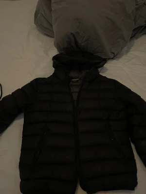 Svart pufferjacka från Colmar - Säljer en svart pufferjacka från Colmar med huva och dragkedja. Jackan har ett stilrent, quiltat mönster och två fickor med dragkedja. Perfekt för kalla dagar och riktigt snygg streetstil. Colmar-logga på ärmen ger en exklusiv touch.