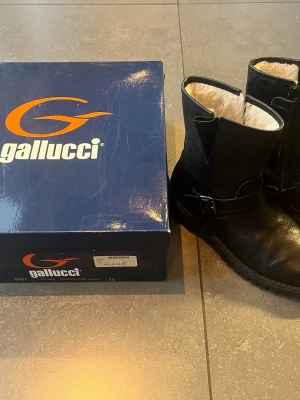 Svarta boots från Gallucci, stl 36 - Säljer ett par svarta skinn boots från Italienska Gallucci i storlek 36. Skorna är i skinn med grov sula, dragkedja på sidan och spänndetalj vid ankeln. Insidan är fodrad med mjukt ljust material för extra komfort. Perfekta för dig som gillar en stilren och robust look.