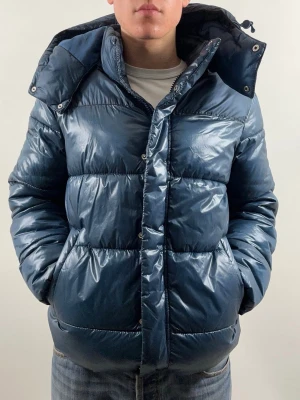 Esprit Pufferjacket Petrol Blue - Snygg blå pufferjacka från Esprit med glansig finish och huva. Jackan har dragkedja och tryckknappar framtill, två sidofickor och quiltad design. Perfekt för kalla dagar och har en modern, loose passform. Materialet är polyester.