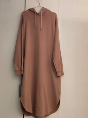 Beige-rosa hoodieklänning från Paulmark - Mysig beige långärmad hoodieklänning från Paulmark i 100% bomull. Klänningen har huva med snörning, ribbade muddar och rundad nederkant. Perfekt för en avslappnad och chill stil. 