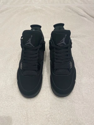 Nike Air Jordan 4 Retro Black  - Säljer ett par helsvarta Nike Air Jordan 4 Retro Black Cat sneakers. Skorna har meshpaneler, mockadetaljer och klassisk snörning. Ikonisk Jumpman-logga på plösen och en chunky sula som ger grym streetstil. Perfekta för dig som vill ha en clean och tidlös look.