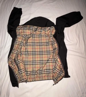 Burberry rutig vindjacka med huva - Säljer en snygg Burberry vindjacka med klassiskt beige rutigt mönster på ena sidan och svart på andra. Jackan är vändbar, har huva, dragkedja och elastiska ärmslut. Perfekt för dig som vill ha en stilren jacka med ikonisk Burberry-look.