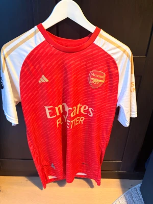 Arsenal Ödegaard tröja Adidas  - Säljer en officiell Arsenal matchtröja från Adidas i storlek M med Ödegaard och nummer 8 på ryggen. Tröjan är röd med vita och guldiga detaljer, har rund hals och korta ärmar. Materialet är lätt polyester och mönstret är diskret randigt. Perfekt för fotbollsfans!