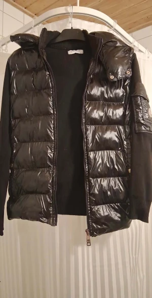 Svart pufferjacka från Moncler - Säljer en svart pufferjacka från Moncler i storlek S. Jackan har glansig quiltad front och detaljer på ärmen med Moncler-logga. Ärmarna är stickade och ger en schysst kontrast mot det blanka materialet. Dragkedja framtill och ribbad krage för extra komfort. Nfc finns på logan