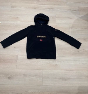 Svart vindjacka från Napapijri - Svart vindjacka från Napapijri med huva och halv dragkedja. Jackan har en stor logga och norsk flagga framtill samt patch på ärmen. Praktisk och stilren design med lång ärm och justerbar nederkant. Kom dm för eventuella frågor  🧐 pris kan diskuteras har använt 2-5 gnr. 
