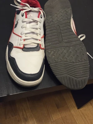 US Polo Assn sneakers vit/röd/blå - Snygga sneakers från US Polo Assn i vit, röd och mörkblå färg. Skorna har snörning, platt sula och rund tå. Märkets logga syns på sidan och insidan är fodrad i rött tyg. Materialet är en mix av syntet och skinn för en sportig vibe.