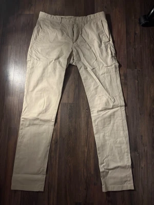 Beige Bowery slim fit chinos - Snygga beige chinos från Bowery med slim fit-passning. Klassisk design med framfickor, bakfickor med knapp och bälteshällor. Tillverkade i mjuk bomull, perfekta för en clean och stilren look. Dragkedja i gylfen och diskreta sömmar.