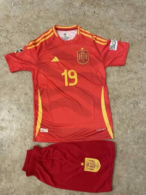 Spaniens Lamine Yamal fotbollsset - Säljer ett rött fotbollsset från Adidas med Spaniens landslagströja och shorts, nummer 19 och Lamine Yamal på ryggen. Tröjan har gula detaljer och emblem, samt tryck på ärmarna. Shortsen är röda med gul badge. Perfekt för dig som älskar fotboll och Spanien.