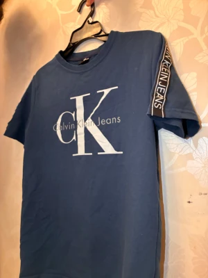 Blå Calvin Klein Jeans t-shirt - Snygg blå t-shirt från Calvin Klein Jeans med stort CK-tryck i vitt på bröstet och logoband på ärmarna. Klassisk rund hals och korta ärmar. Perfekt för dig som gillar stilrena och trendiga märkesplagg. Storlek kan vara från XS - M. Priset kan diskuteras!