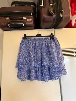 Blå volangkjol från Odd Molly - Supersöt blå kortkjol från Odd Molly med volanger och elastisk midja. Kjolen har ett detaljerat blommigt mönster i vitt och blått och är tillverkad i ett lätt och luftigt material, perfekt för varma dagar. Passformen är normal och kjolen har en feminin och flowy känsla. Från märket Odd Molly. Storlek 2 som enligt deras storleksguide motsvarar en M. Mycket fint skick, se sista bild på resonen i midjan. Den kanske behöver fixas till lite. Kan kan vara något ojämn. 