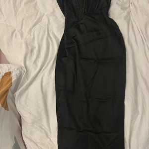 Svart långklänning från Boohoo - Supersnygg svart långklänning från Boohoo med axelbandslös design och markerad byst. Klänningen har en figurnära passform, slät satinliknande yta och dold dragkedja bak. Perfekt för dig som vill ha en elegant och stilren look. Klänningen har ett slit på baksidan.