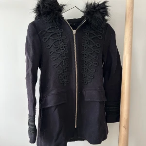 Svart kappa med fuskpäls från Zara - Svart kappa från Zara Woman med snygga dekorativa band framtill och dragkedja. Stor huva med fluffig fuskpäls och två fejk fickor. Ärmarna har detaljer och kappan är i ett mjukt ull-liknande material. Passar som S.