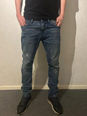 Tom Tailor blå slim jeans - Säljer ett par blå jeans från Tom Tailor, modell Piers Slim. Jeansen har en snygg tvättad look med lätt slitna detaljer och klassiska bakfickor. De är i slim fit och har normal midja. 