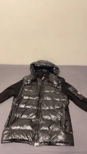 Svart duncardigan från Moncler med luva - Pris kan diskuteras‼️‼️Svart dunväst från Moncler med glansig yta och stickade svarta ärmar. Cardigan har en avtagbar luva, dragkedja framtill och Moncler-logga på ärmen. Quiltad design och praktiska fickor med knappstängning. Perfekt för dig som vill ha en snygg och varm look.