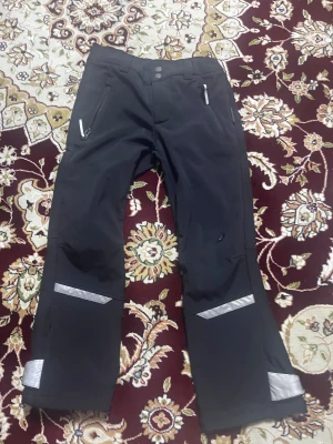 Svarta täckbyxor med reflexdetaljer - Svarta täckbyxor från Didriksons med reflexdetaljer på benen, dragkedjefickor och dubbla knappar i midjan. Byxorna har justerbara hängslen och är perfekta för kalla dagar. Materialet är slitstarkt och vattenavvisande syntet. Ett litet håll i 