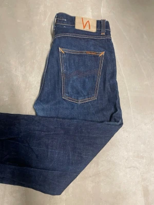 Mörkblå jeans från Nudie Jeans Co - Steady Eddie - Snygga mörkblå jeans från Nudie Jeans Co med klassisk femficksdesign och kontrastsömmar i orange. Modellen har rak passform och normal midja, tillverkade i slitstarkt jeanstyg. Perfekta för dig som gillar stilrena och tidlösa jeans.