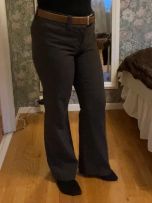 Mörkbruna kostymbyxor med bootcut - Snygga mörkbruna kostymbyxor med svagt rutigt mönster och bootcut-ben. Byxorna har hög midja, bälteshällor och stängs med både hake och dragkedja. Perfekt för dig som gillar klassisk stil med en modern twist.