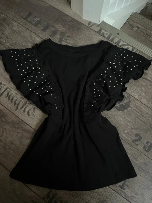 Svart topp med volang och pärlor - Snygg svart topp med korta volangärmar dekorerade med små vita pärlor. Toppen har en rund hals och är tillverkad i mjukt tyg som sitter bekvämt. Perfekt för dig som gillar detaljer och vill sticka ut lite extra med coola volanger och pärlor.
