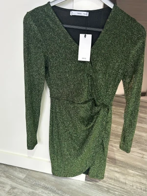 Grön glittrig kortklänning från MNG - Supersnygg grön glittrig kortklänning från MNG med v-ringning och långärmad design. Klänningen har omlott-effekt framtill och en liten slits vid nederkanten. Perfekt för dig som vill sticka ut med lite extra glitter och cool passform.