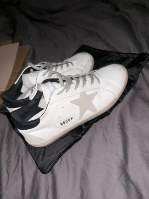 Golden Goose vita sneakers med stjärna - Golden Goose sneakers i vitt skinn med grå mockadetaljer och en ikonisk stjärna på sidan. Skorna har grå snörning, svart häl och rund tå. Medföljer originalkartong och dustbag. Perfekta för dig som gillar stilrena och trendiga sneakers.