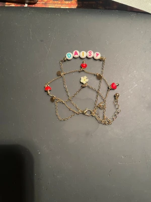 Guldig kedjeväst med svamp- och blomdetaljer - Guldig kedjeväst med flera lager och detaljer som röda svampberlocker, små guldplattor och en söt blomma. Ena kedjan har pärlor med bokstäverna "Daisy" i olika färger.